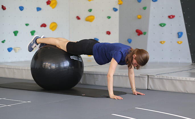 Eine Mitarbeiterin der TheraMedis Lahn Dill macht Übungen auf einem Pezziball vor der Boulder-Wand